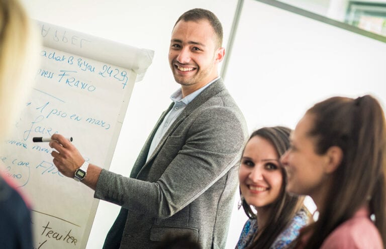 Un groupe de personne d'âges et de nationalités différentes sont rassemblées en réunion devant un paperboard