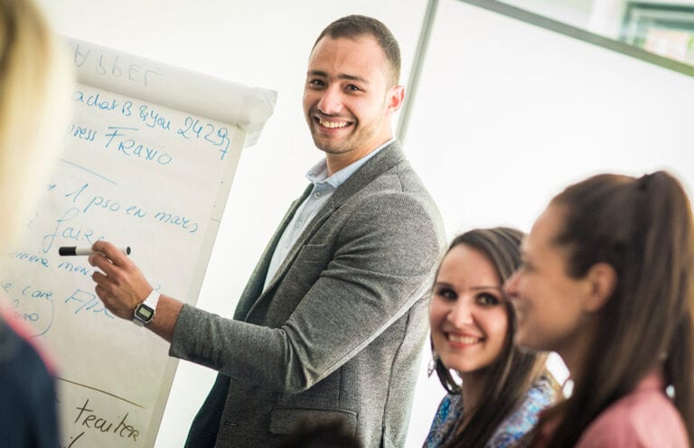 Un groupe de personne d'âges et de nationalités différentes sont rassemblées en réunion devant un paperboard