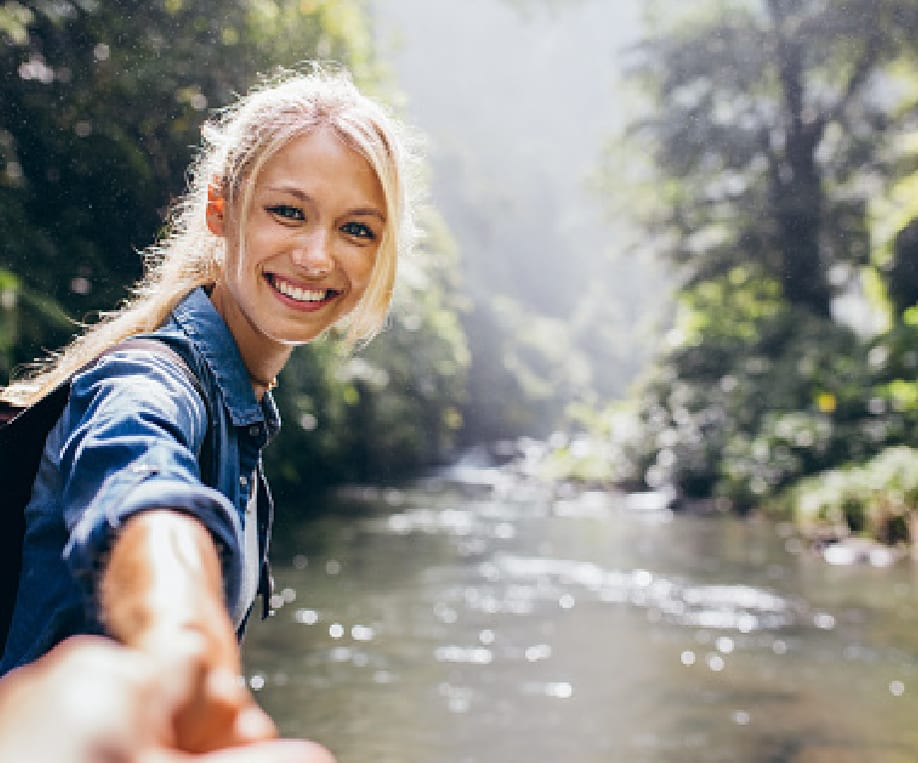 Une jeune femme en tend la main en souriant à une autre personne dont on ne voit que la main au premier plan. En arrière plan il y a une petite rivière bordée d'arbre. On peut donc penser qu'elle aide l'autre personne à traverser.