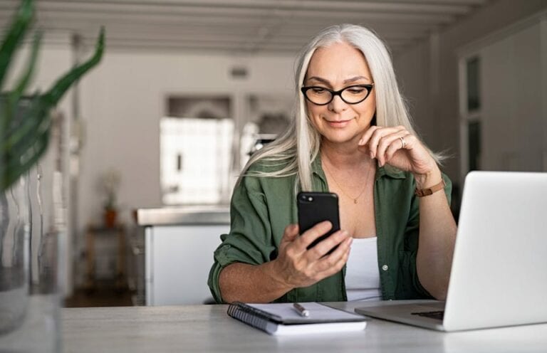 Femme agée assise à la table de son salon, elle regarde son téléphone en souriant